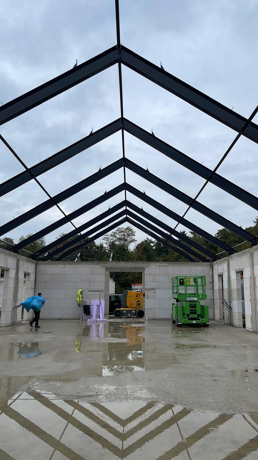 Rohbau einer Stahlhalle mit schwarzem Dachtragwerk, Betonwänden, grüner Hebebühne und Arbeiter auf nassem Betonboden